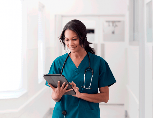 mulher preta veste roupa de hospital azul, tem estetoscópio no pescoço, segura tablet enquanto o olha; ao fundo vemos detalhes de um hospital em branco.