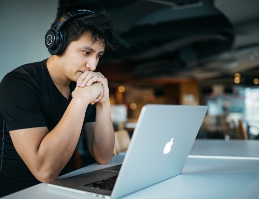 Homem branco de cabelos escuros e camiseta preta está sentado em frente a um notebook da apple, ele usa fones de ouvido pretos e tem as duas mãos abaixo do queixo em feição cencentrada.