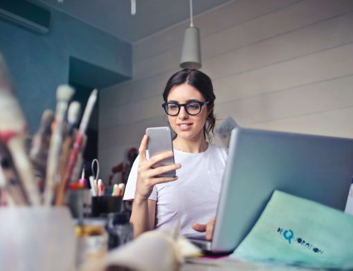 mulher jovem tem cabelos escuros, usa óculos preto e camiseta branca, olha para um celular cinza; ao seu redor há um notebook, papeis, pincéis e itens de pintura.