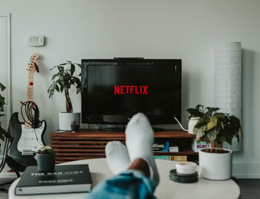 foto de sala de estar com televisão com símbolo da netflix; rack de madeira, vasos de plantas, uma guitarra e uma mesa redonda branca com vasos e livros e dois pés apoiados de meia.