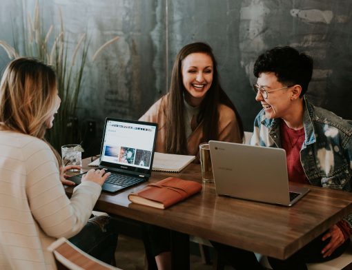 mulher de cabelo loiro e blusa bege, mexe no computador de um lado da mesa quanto um homem e uma mulher do outro lado sorriem; há um notebook e papeis do seu lado.