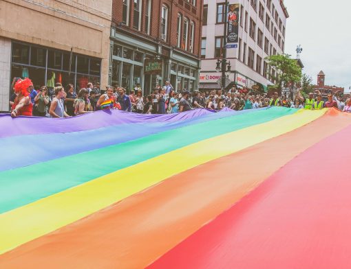 no primeiro plano, uma bandeira do arco iris representando o orgulho LGBT está esticada sendo segurada por várias pessoas no segundo plano do lado esquerdo; ao fundo vemos prédios.