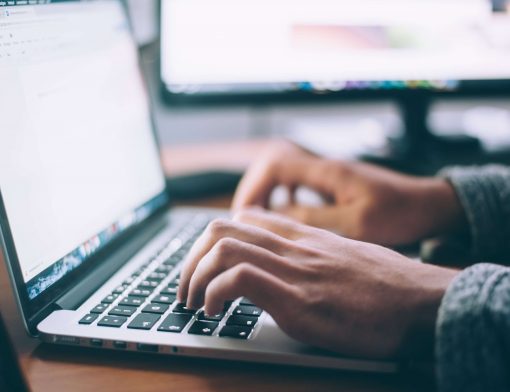 foco em mãos masculinas digitando em um notebook cinza; ao fundo vemos uma parte de um monitor ligado.