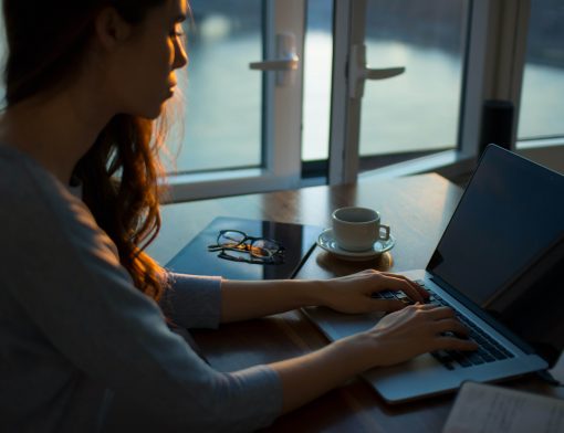 imagem com uma luz escura de uma mulher de perfil escrevendo em um notebook; ao lado dela, na mesa é possível ver um tablet, um óculos e uma xícara.