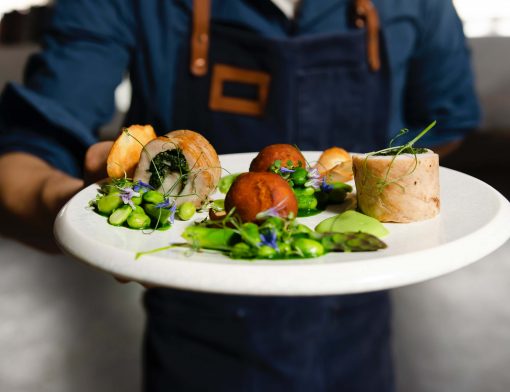 foco em um prato branco com alimentos verdes, vermelhos e bege segurado por um homem de camisa azul e avental, onde vemos apenas o tronco.