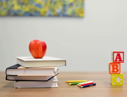 imagem de uma mesa com livros empilhados e uma maça no topo, com lápis coloridos ao lado e três cubos com escritos ABC.