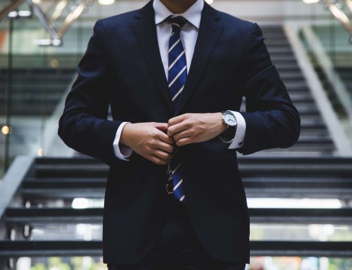 foco no tronco de um homem de terno azul, gravata azul com listras cinzas, ele está fechando os botões e usa um relógio preto.