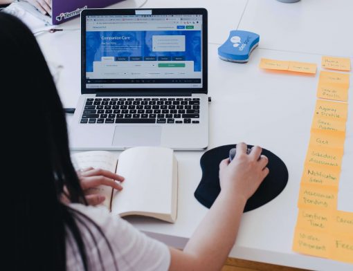 foco em uma mulher de cabelo preto solto, de costas mexe em um mouse sob a mesa, ela olha um notebook; ao lado há vários post its, um livro aberto e outros itens.