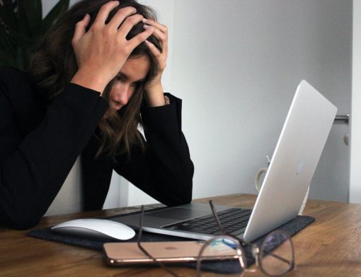 mulher veste casaco preto, tem as mãos na cabeça em sinal de preocupação. Ela olha notebook sob uma mesa que tem um óculos, celular e mouse ao lado.