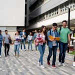 quatro jovens entre homens e mulheres estao do lado esquerdo no primeiro plano andando, conversando e olhando livros juntos; ao fundo é possível, ver mais estudantes e um prédio.