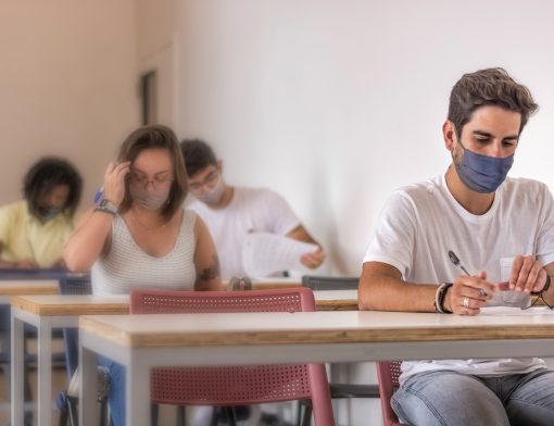 imagem de uma sala de aula onde jovens diversos usam máscara e estão olhando provas em suas carteiras.