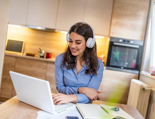 mulher branca de cabelos escuros usa camisa azul e fones de ouvido brancos; uma de suas mãos está sob um notebook branco repousado na mesa, onde também vemos livros; ao fundo vemos armários e decoração de uma cozinha.