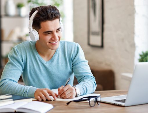 homem usa camiseta azul e fone branco, escreve em papel enquanto olha notebook sob a mesa, que tem outro livro aberto; ao fundo vemos quadro em uma parede.