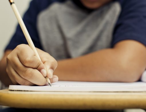 foco nos braços de um jovem apoiado em uma carteira escolar escrevendo em um papel.