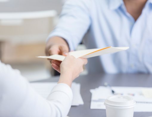 imagem com foco em um homem oferecendo um papel a uma mulher que está recebendo; é possível ver outros papeis e um copo de café na mesa abaixo.