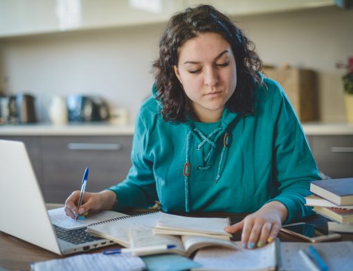 imagem de uma mulher de cabelos cursos escuros, que usa blusa verde e olha papeis em cima da mesa com um computador do lado; ao fundo vemos armário.