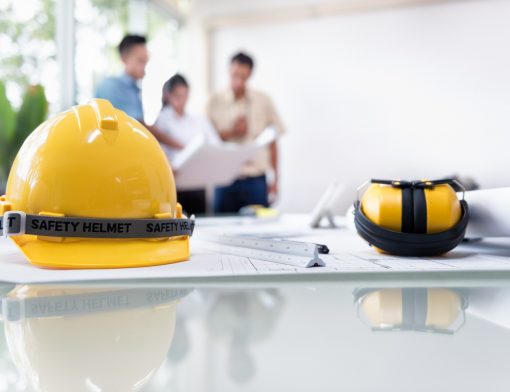 imagem com foco em uma mesa de vidro com chapeu e fones de segurança em cima junto com papeis; ao fundo vemos uma mulher e dois homens olhando um projeto em papel.