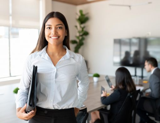 mulher branca, com cabelo moreno. Tem expressão feliz, sorri enquanto segura uma pasta embaixo do seu braço direito; ela usa camisa branca listrada de azul; ao fundo é possível ver uma mesa de reuniões com duas pessoas sentadas e uma televisão na parede