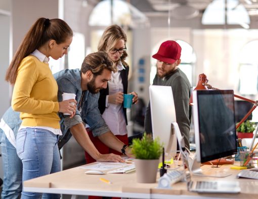 quatro pessoas diferentes estão ao redor de uma mesa olhando um monitor, algumas de pé e outras inclinadas ou sentadas na mesa; vemos teclado, vaso de plantas e outros itens de escritório ao lado.