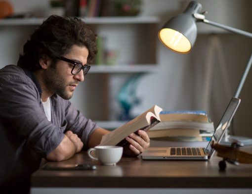 foto de perfil de um homem lendo um livro apoiado em uma mesa que tem um notebook e outros livros empilhados; vemos um abajur ao lado e prateleiras brancas ao fundo.