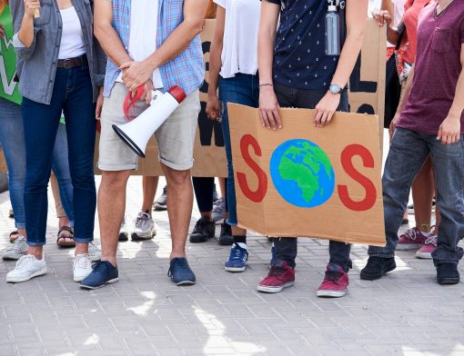 imagem de várias pessoas de pé na altura do tronco; uma do primeiro plano segura um megafone e ao seu lado uma pessoa segura uma placa com o escrito SOS sendo a letra O, o símbolo do planeta terra.