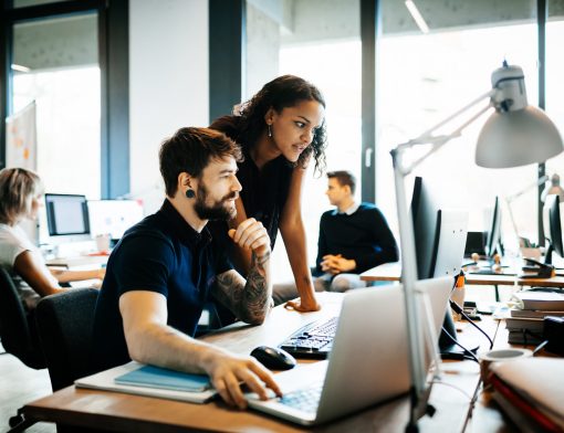 um homem de barba, camisa azul e várias tatuagens está setado ao redor de uma mesa, digitando num notebook e olhando para um monitor. Ao lado dele há uma mulher preta apoiada na mesa olhando também o monitor; ao fundo vemos um homem sentado de perfil.