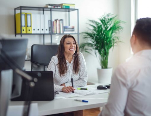 mulher branca de cabelo castanho longo, veste camisa branca, está atrás de uma mesa olhando para um homem que está na frente da mesa, usando camisa branca; ela sorri e segura uma caneta; na mesa é possível ver papeis e canetas e ao redor plantas, cadeira, estante e livros.