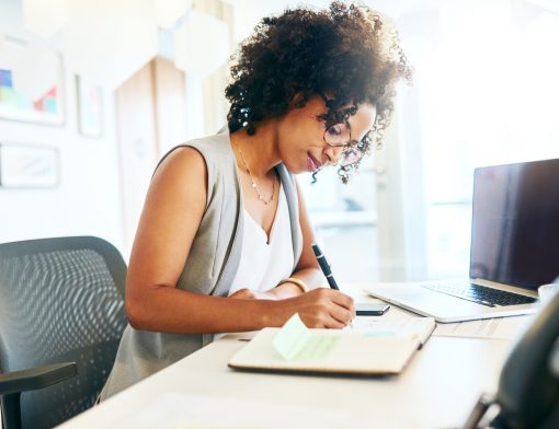 mulher preta de cabelo cacheado, usa regata branca e colete cinza; ela está de lado com a cabeça inclinada olhando o papel em que está escrevendo em cima de uma mesa; é possível ver um notebook do seu lado.