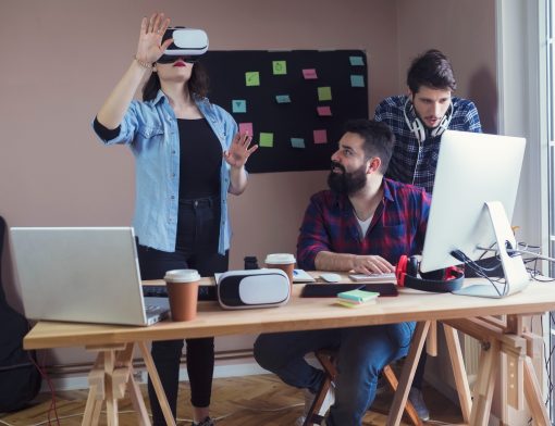 mesa com computadores e óculos de realidade virtual; em pé uma mulher usa um dos óculos, está com as mãos levantadas; ao lado dela, um homem observa enquanto outro olha o computador.