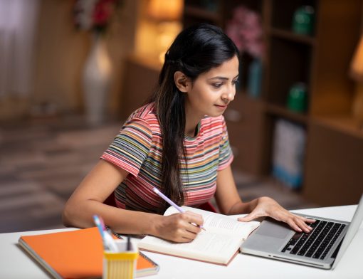 imagem de mulher de cabelos pretos longos olhando um computador; ela veste blusa listrada de vermelho e verde, tem uma mesa no notebook e outra escrevendo em um livro.