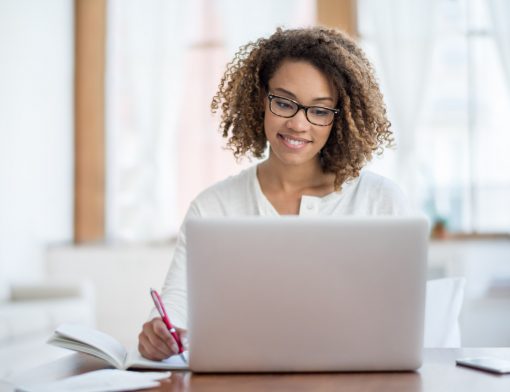 mulher preta de cabelo cacheado curto usa blusa branca escreve em um papel enquanto olha para notebook branco.