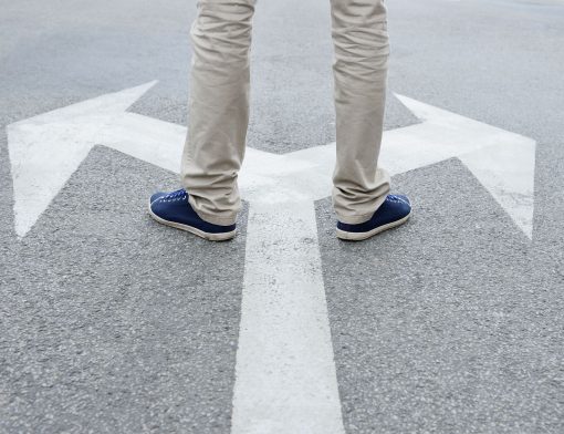 foco nas pernas de homem que veste calça cinza e tenis azul; ele está em uma rua pisando acima de um y com setas mostrando caminhos.