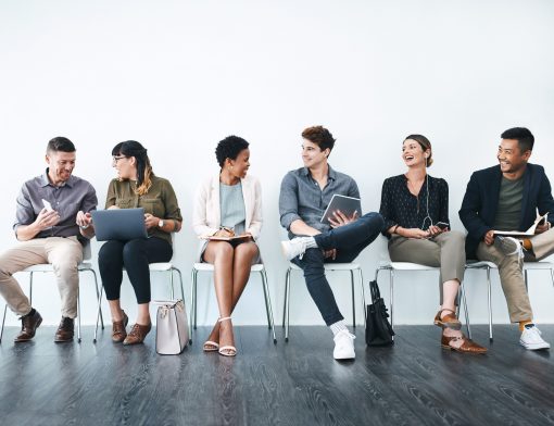 parede branca e chão cinza, com seis pessoas diversas entre homens e mulheres de etnias diferentes estão sentadas em cadeiras; eles se olham em tom de conversa.