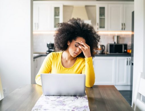 mulher preta de cabelo crespo usa blusa amarela; ela está com os olhos fechados e a mão no rosto; ao redor há uma mesa com notebook e ao fundo uma cozinha com móveis brancos.