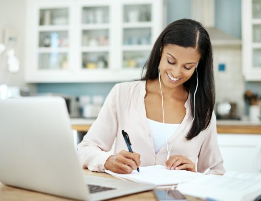 mulher preta tem cabelos lisos, está com fone de ouvido, tem camisa branca, escreve em um papel; sob a mesa há um notebook e outros papeis; ao fundo vemos armário de cozinha branco.