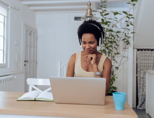 mulher preta de cabelo black usa fones de ouvido e regata bege, tem uma mão no queixo e olha notebook sob uma mesa,. que tem um copo azul e um livro ao lado; ao fundo vemos porta branca e planta.