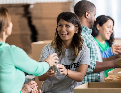 uma mulher de camiseta cinza entrega um item para uma mulher de blusa verde que está de costas; ao fundo vemos outras pessoas manipulando caixas e caixas fechadas atrás.