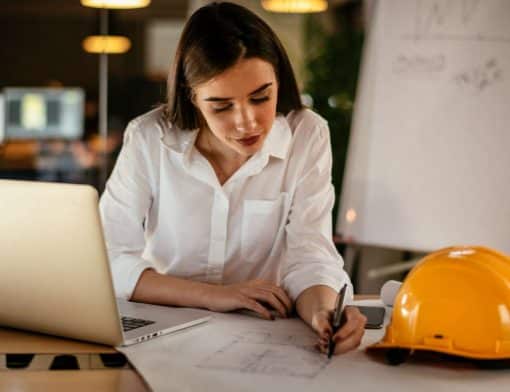 imagem de uma mulher escrevendo em alguns papeis de engenharia, ela veste camisa branca; ao lado dela tem um chapeu de construção e um notebook na mesa.