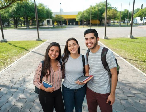 três jovens estão no primeiro plano, duas mulheres e um homem na esquerda; as mulheres seguram livros e todos usam mochilas; ao fundo é possível ver calçada, grama e um prédio grande.