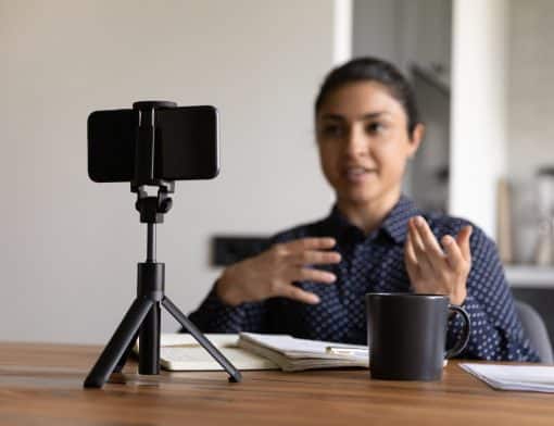 imagem de mulher ao fundo gesticulando na frente da câmera de celular que está no primeiro plano. Na mesa na frente dela existem papeis e uma caneca.