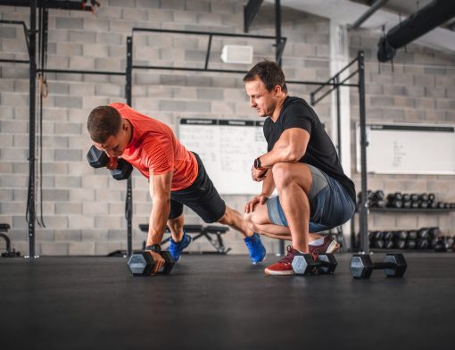 imagem de uma academia ao fundo, um homem está na direita ajoelhado olhando um outro homem que está fazendo um exercício na horizontal se levantando com pesos nas mãos.