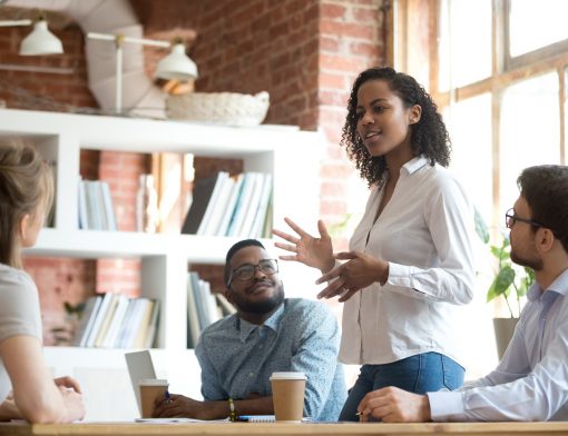 uma mulher preta de cabelos cacheados curtos, veste camisa branca e calça jeans, está gesticulando com as mãos enquanto outros 3 colegas sentados ao redor da mesa olham para ela; vemos notebook e copos de café na mesa e uma estante branca com livros ao fundo.