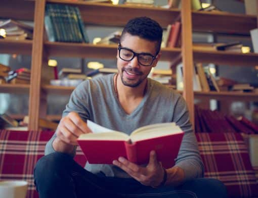 homem preto usa óculos, camiseta de manga comprida cinza e calça escura; ele sorri enquanto segura um livro aberto de capa vermelha; está sentado em sofá xadrez vermelho e ao fundo vemos estante com livros.