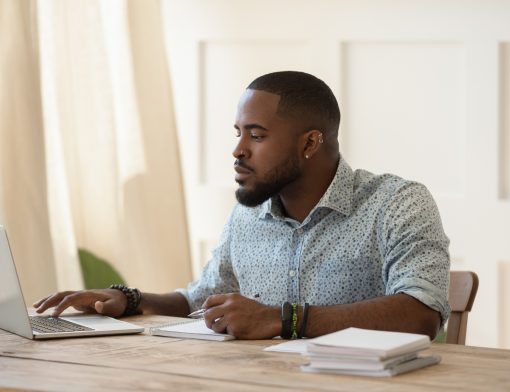 homem preto usa camisa azul de bolinhas pretas, olha um notebook sob uma mesa com papeis.