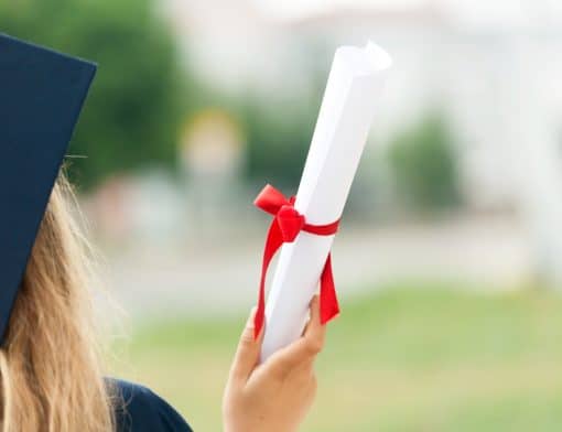 foto de uma parte de da cabeça de uma mulher de costas que veste roupa e chapeu de formatura e segura diploma levantado.