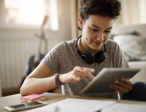 imagem de uma mulher branca de cabelos pretos curtos, ela veste camiseta cinza e um headphone no pescoço enquanto segura um tablet; é possível ver também uma mesa com papeis desfocadas e o fundo de um quarto.