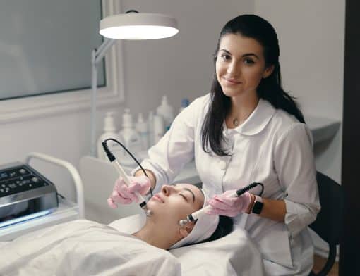 mulher de cabelo preto e jaleco branco, sorri pra foto, usa luvas cor de rosa e segura equipamentos encostados no rosto de outra mulher que está deitada em uma maca; ao redor vemos uma luminária e outros equipamentos médicos.