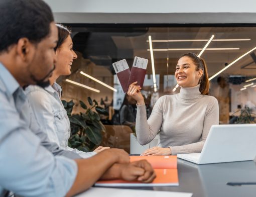 uma mulher de cabelos presos está sentada, segura dois passaportes; na sua frente há uma mulher branca e um homem preto olhando a primeira mulher; na mesa vemos livro laranja e notebook branco.