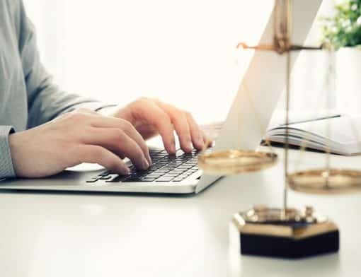 mesa branca com um notebook e um homem com mãos no teclado; ao lado vemos livro aberto, um vaso de plantas e a balança simbolo do equilíbrio.