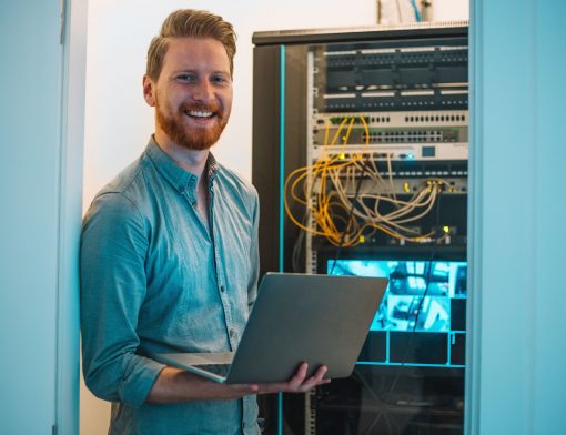 homem ruivo de pé, usa camisa cinza e segura notebook, ele sorri olhando para a foto e atrás dele há uma torre eletronica com fios e imagens de cameras.